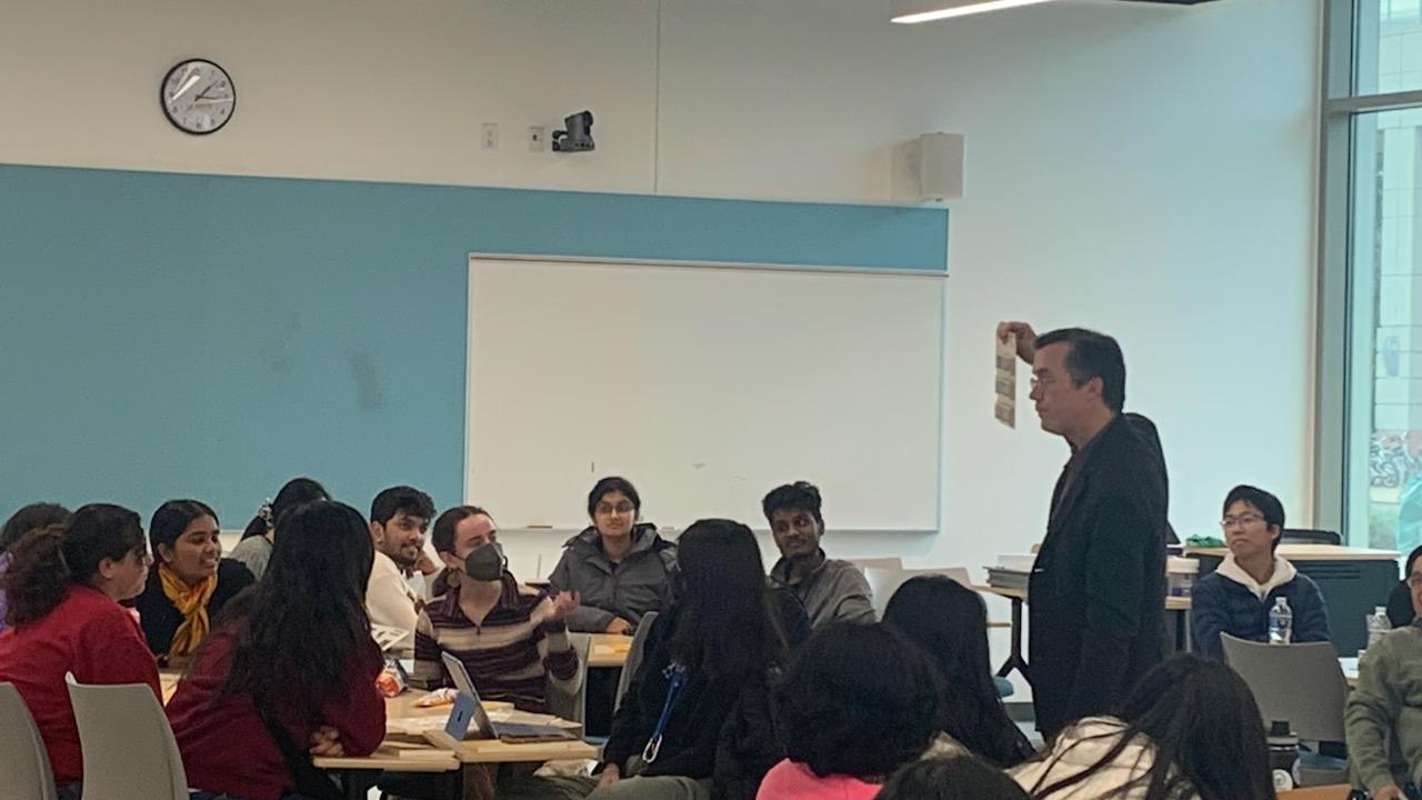 Multiple students sitting in a classroom with some students watching a faculty member hold up a paper.