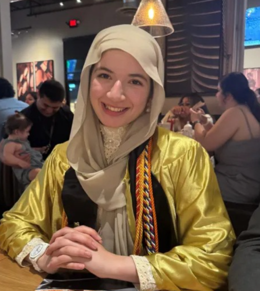 Smiling woman in beige hijab and gold graduation gown with cords at a restaurant, photo