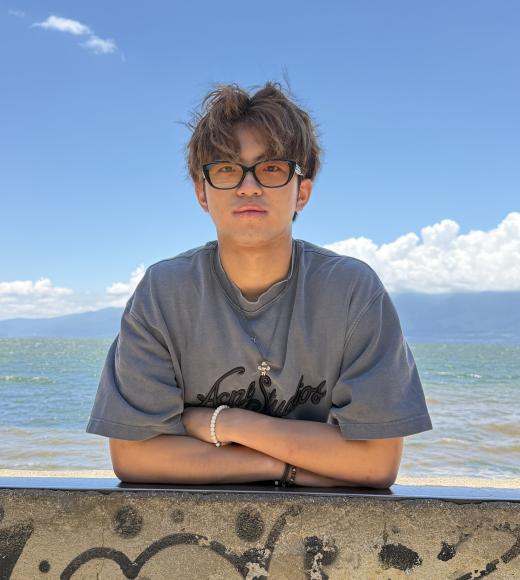 Young man wearing glasses and gray tee leaning on seaside wall, mountains and blue sky (photo)