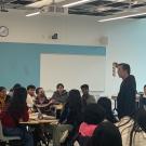 Multiple students sitting in a classroom with some students watching a faculty member hold up a paper.